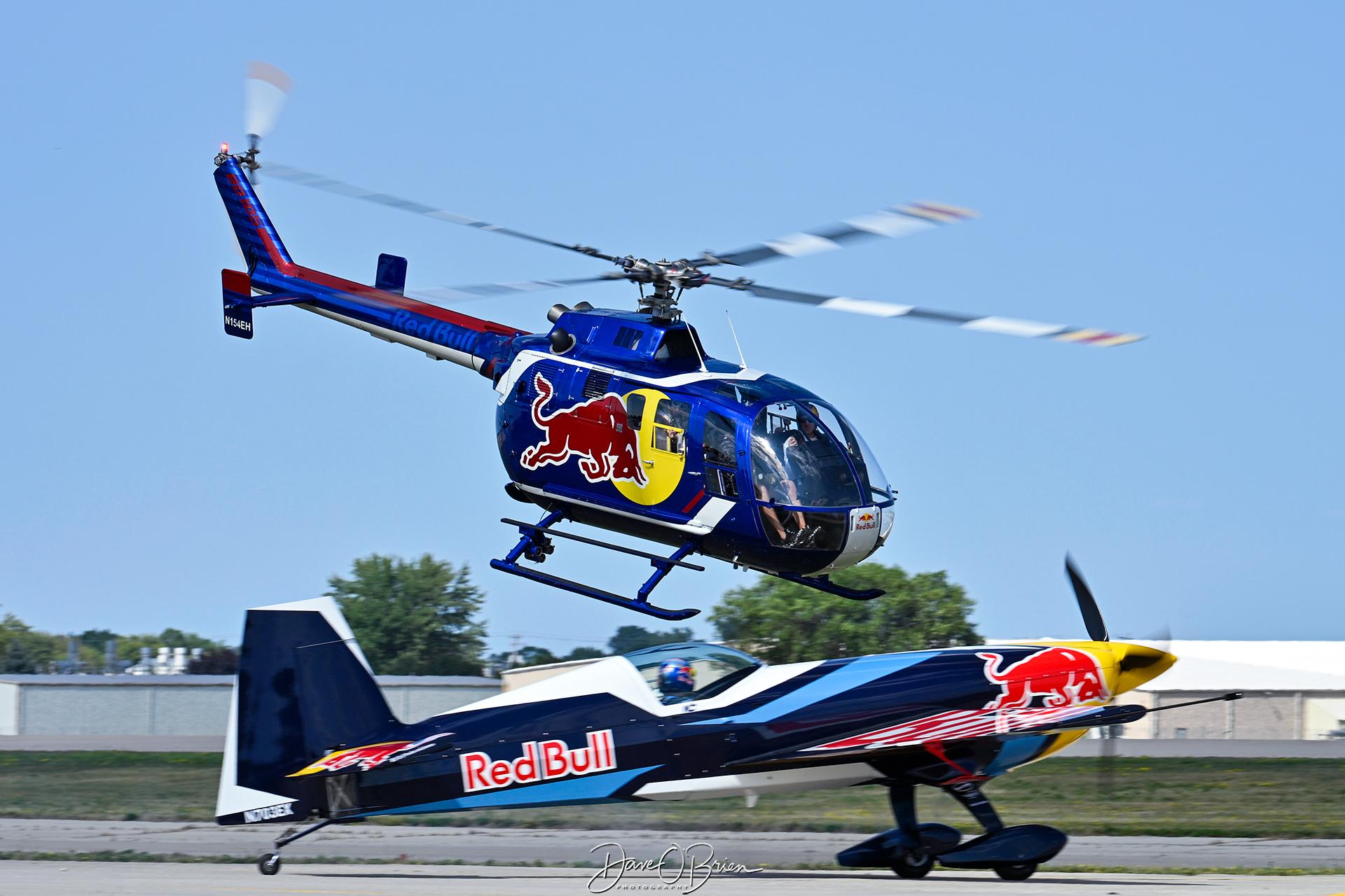 Aircraft N154EH, Red Bull helo taking off while the Red Bull Extra holds on the taxiway, at KOSH, photo by topgunphotography